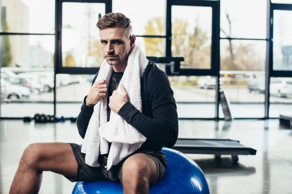 Schöner Sportler sitzt mit Handtuch auf Fitnessball und schaut in die Kamera im Fitnessstudio — Stockfoto