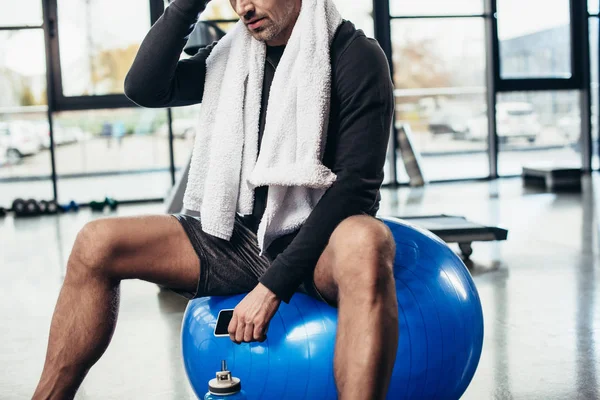 Imagem cortada de desportista sentado na bola de fitness com toalha e smartphone no ginásio — Fotografia de Stock