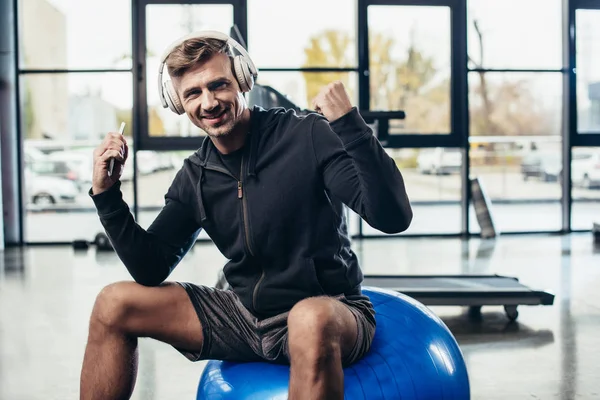 Deportista guapo feliz sentado en la pelota de fitness y escuchar música en el gimnasio - foto de stock