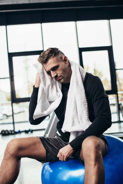 Schöner müder Sportler sitzt mit Handtuch auf Fitnessball im Fitnessstudio — Stockfoto