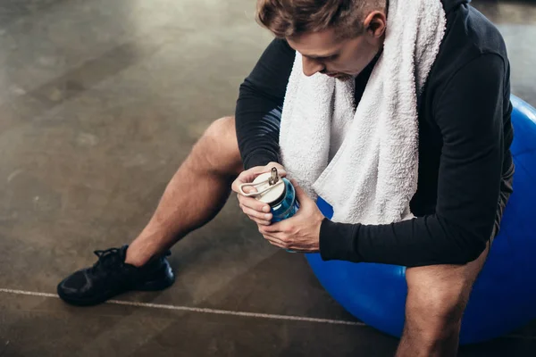 Vista de alto ângulo de desportista cansado bonito sentado na bola de fitness com toalha e garrafa de esporte no ginásio — Fotografia de Stock