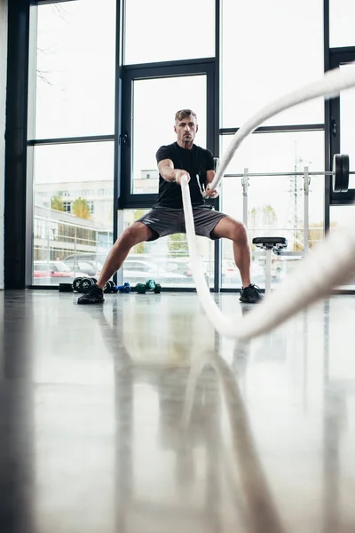 Selective focus of handsome sportsman working out with ropes in gym — Stock Photo
