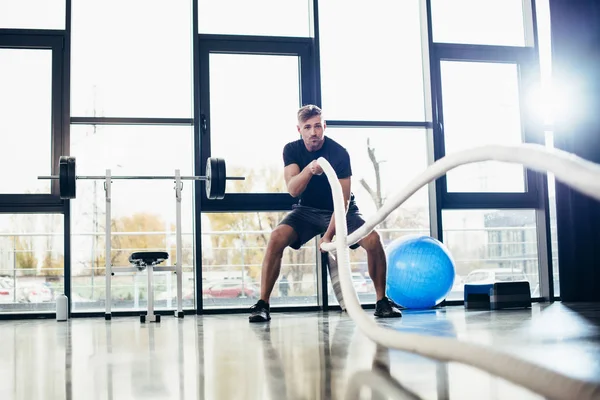 Full length of handsome sportsman training with ropes in gym — Stock Photo