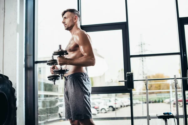 Vue latérale du beau sportif torse nu s'entraînant avec des haltères dans la salle de gym — Photo de stock