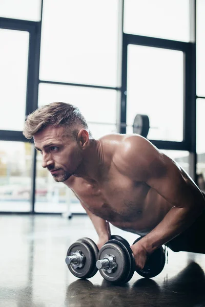 Portrait of handsome shirtless sportsman doing plank on dumbbells in gym — Stock Photo