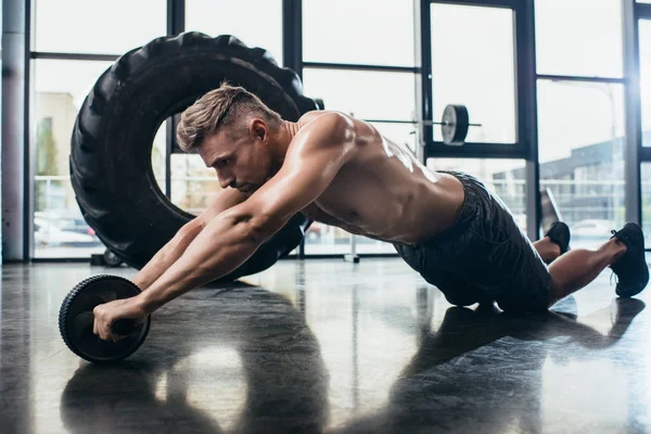 Handsome shirtless muscular sportsman exercising with abs roller in gym — Stock Photo