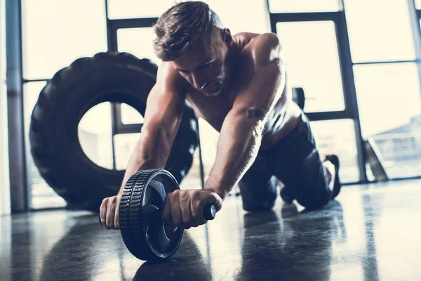 Beau sportif sans chemise athlétique faisant de l'exercice avec un rouleau abdominal dans la salle de gym — Photo de stock