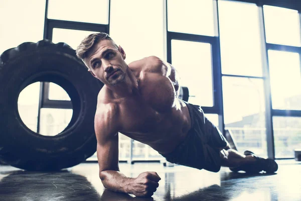 Handsome shirtless sportsman doing plank on one hand in gym — Stock Photo