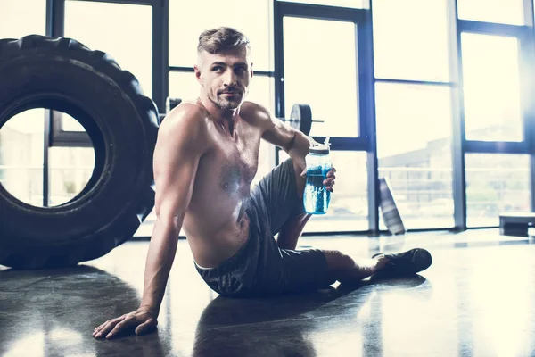 Beau sportif torse nu assis sur le sol dans la salle de gym et tenant bouteille de sport — Photo de stock
