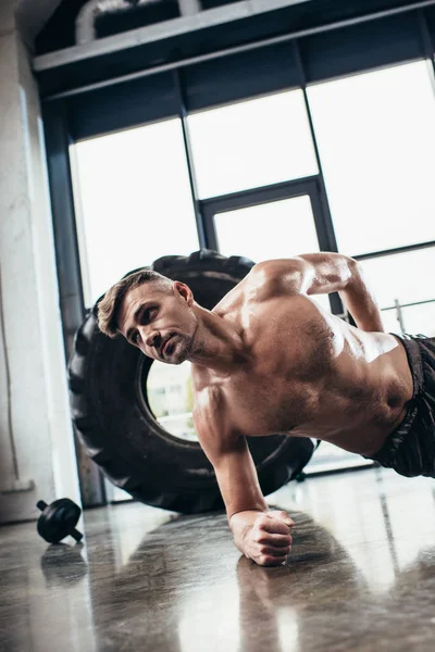 Handsome shirtless muscular sportsman doing plank on one hand in gym — Stock Photo