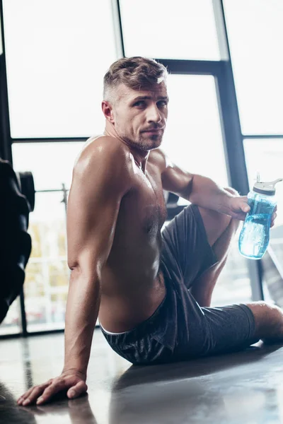 Handsome shirtless sportsman sitting on floor in gym and holding sport bottle, looking at camera — Stock Photo