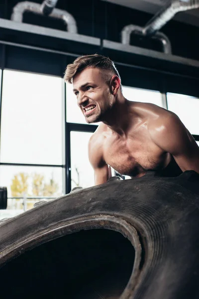 Apuesto deportista sin camisa levantando neumáticos y haciendo muecas en el gimnasio - foto de stock