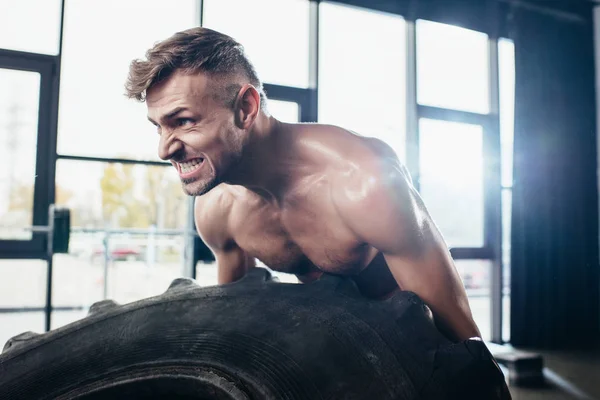 Handsome muscular sportsman lifting tire and grimacing in gym — Stock Photo