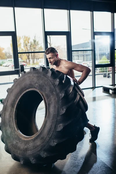 Handsome shirtless sportsman lifting tire in sport center — Stock Photo