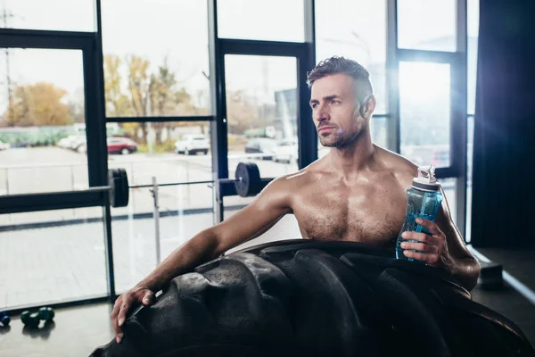 Apuesto deportista sin camisa apoyado en el neumático y la celebración de la botella de deporte en el gimnasio — Stock Photo