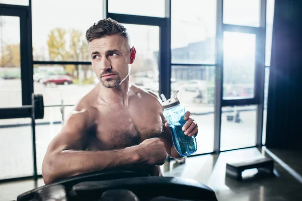 Apuesto deportista sin camisa apoyado en el neumático y la celebración de la botella de deporte en el gimnasio, mirando hacia otro lado - foto de stock