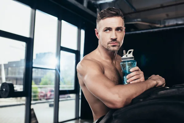 Apuesto sin camisa deportista muscular apoyado en el neumático y la celebración de la botella de deporte en el gimnasio — Stock Photo