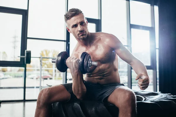 Handsome shirtless sportsman sitting on tire and exercising with in gym dumbbell — Stock Photo