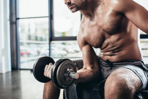 Cropped image of sweaty shirtless sportsman sitting on tire and exercising with in gym dumbbell — Stock Photo