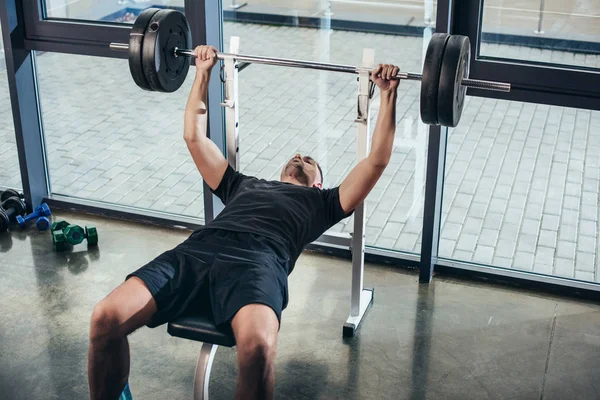 Hombre deportivo guapo levantando la barra con pesas mientras está acostado en el banco en el gimnasio - foto de stock