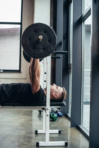 Seitenansicht eines gut aussehenden, athletischen Mannes, der eine Langhantel mit Gewichten hebt, während er im Fitnessstudio auf einer Bank liegt — Stockfoto