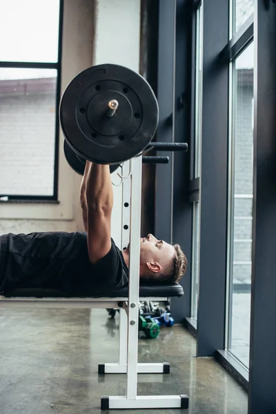 Vista lateral do homem atlético bonito levantando barbell com pesos enquanto deitado no banco no ginásio — Fotografia de Stock