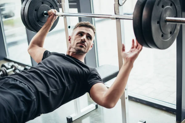 Schöner Sportler auf Bank liegend, um Langhantel in Turnhalle zu heben — Stockfoto