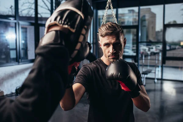 Selektiver Fokus des gutaussehenden Boxers beim Training mit dem Trainer im Fitnessstudio — Stockfoto