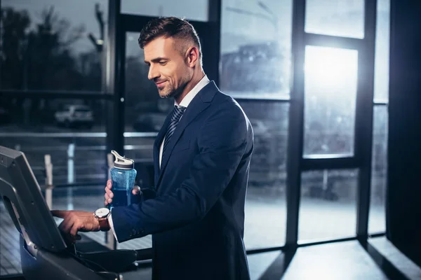 Красивый бизнесмен в костюме тренируется на беговой дорожке и держит спортивную бутылку в тренажерном зале — стоковое фото