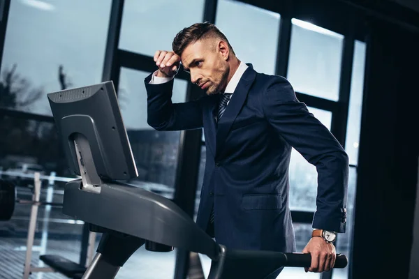 Vista lateral de hombre de negocios guapo cansado en traje haciendo ejercicio en la cinta de correr en el gimnasio - foto de stock