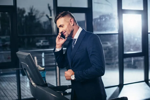 Красивый бизнесмен в костюме тренируется на беговой дорожке и разговаривает по смартфону в тренажерном зале — стоковое фото