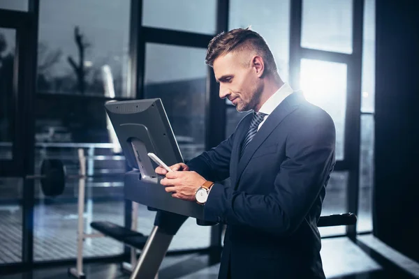Vista lateral del hombre de negocios guapo en traje apoyado en la cinta de correr y el uso de teléfono inteligente en el gimnasio — Stock Photo