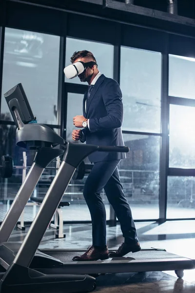 Vue latérale de l'homme d'affaires en costume et casque de réalité virtuelle exercice sur tapis roulant dans la salle de gym — Photo de stock