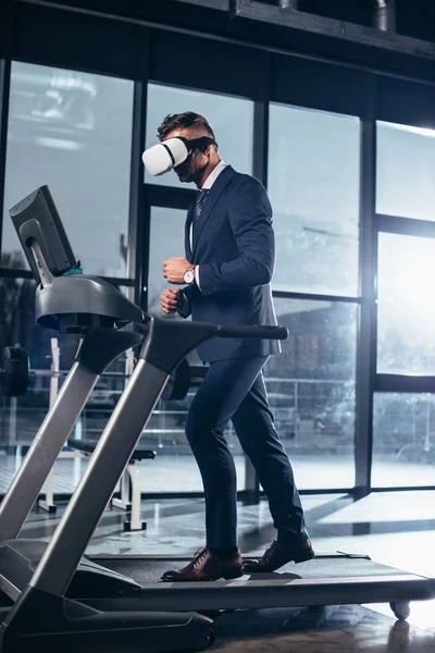 Vista lateral do homem de negócios em terno e fone de ouvido realidade virtual exercendo na esteira na academia — Stock Photo
