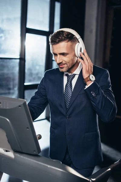 Bel homme d'affaires en costume exercice sur tapis roulant et écouter de la musique avec écouteurs dans la salle de gym — Photo de stock