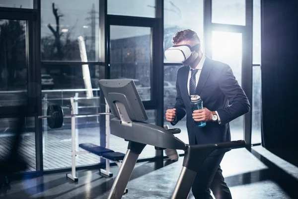 Businessman in suit and virtual reality headset exercising on treadmill and holding sport bottle in gym — Stock Photo