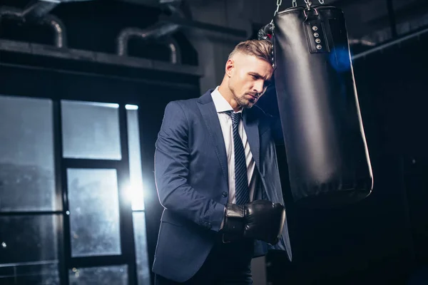 Handsome tired businessman in suit and boxing gloves leaning on punching bag in gym — Stock Photo