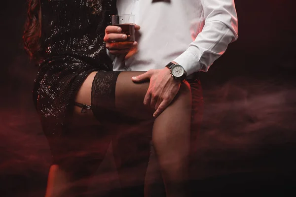 Vista ritagliata della donna erotica che abbraccia l'uomo con un bicchiere di whisky in camera fumosa rossa — Foto stock