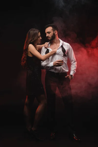 Apasionada mujer abrazo hombre con vaso de whisky en rojo ahumado habitación - foto de stock