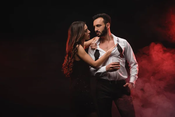 Sensual girl hugging man with glass of whiskey in red smoky room — Stock Photo