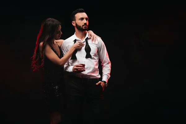 Sexual woman undressing handsome man with glass of whiskey isolated on black — Stock Photo