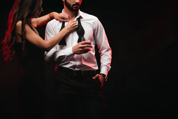 Cropped view of woman undressing man with glass of whiskey isolated on black — Stock Photo
