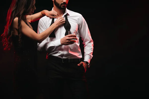 Vue recadrée de femme déshabiller l'homme avec un verre de whisky isolé sur noir — Photo de stock