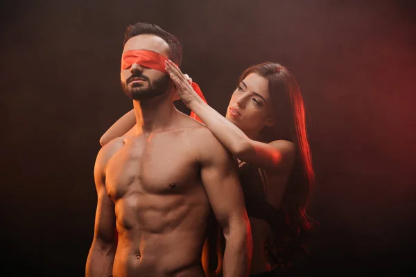 Sexy girl closing eyes of shirtless boyfriend with red ribbon in smoky room — Stock Photo