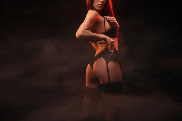 Cropped view of sexy young woman in black lingerie posing in dark smoky room — Stock Photo