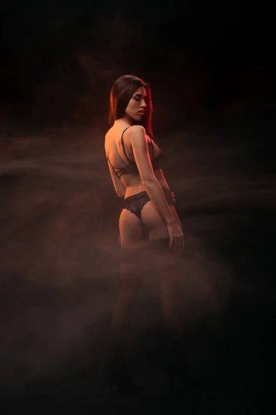 Beautiful sensual young woman in black lingerie posing in dark smoky room — Stock Photo