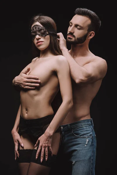 Novio guapo abrazando sexy novia desnuda en máscara de encaje, aislado en negro - foto de stock