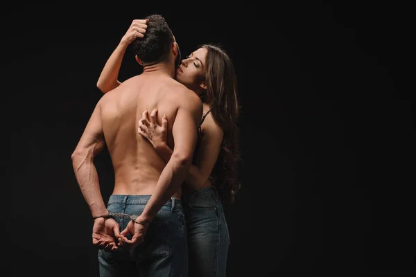 Seductive young woman hugging shirtless man in handcuffs isolated on black — Stock Photo