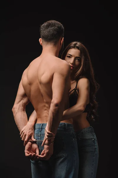 Schöne sexy Mädchen umarmt hemdlosen Mann in Handschellen isoliert auf schwarz — Stockfoto