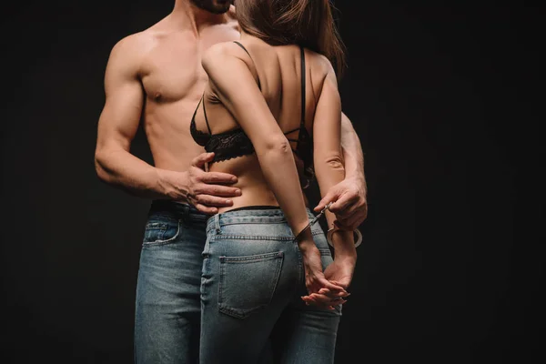 Recortado vista de sin camisa hombre abrazando sexy mujer en esposas aislado en negro - foto de stock
